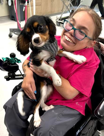 Kayla Milewski smiling in her wheelchair while cradling a Cavalier King Charles spaniel